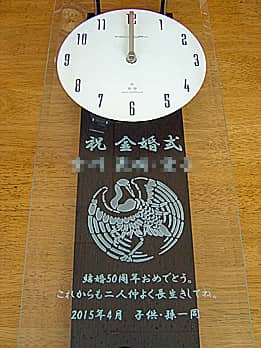 「祝金婚式、両親の名前、鶴のイラスト」を前面ガラスに彫刻した、金婚式のお祝い品用の掛け時計