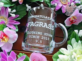 「Congratulations、店名とキャッチフレーズ」を側面に彫刻した、開店祝い用のガラス製マグカップ