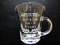 「勤続20周年記念、永年勤続者名、日付、会社名」を彫刻した、永年勤続表彰用のガラス製ティーカップ